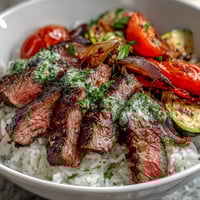 Sliced grilled steak rests atop fluffy rice and roasted veggies in this vibrant Grilled Steak Bowl, drizzled with bright green chimichurri sauce for a hearty meal.